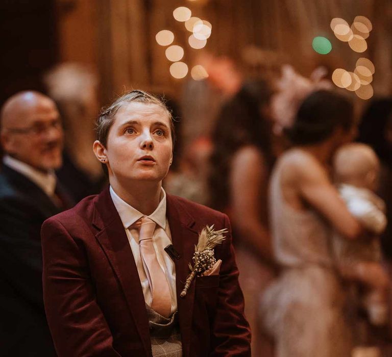 Bride in maroon bridal suit with grey waistcoat, dusky pink tie and autumnal dried flower boutonniere waiting at the end of the alter 