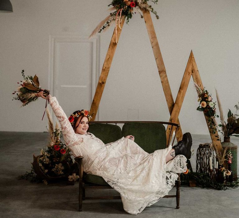 Bride in boho long sleeve 3D applique wedding dress with boho rustic dried flower crown holdingearthy toned garden rose, pampas grass, dried flower and foliage bridal bouquet sitting on crushed velvet olive green loveseat in front of one small and one large wooden triangular wedding arch with pampas grass, foliage, and garden rose arrangements attached, rustic wedding decorations, large plants in vases and pillar candles 