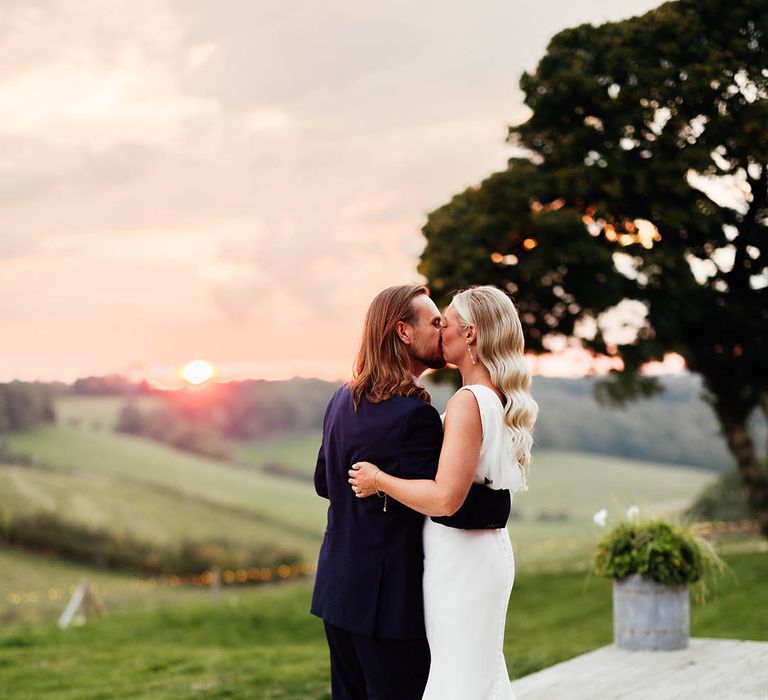 The bride in a Savin London wedding dress and groom in blue suit share a kiss during the sunset over the Surrey Hills 