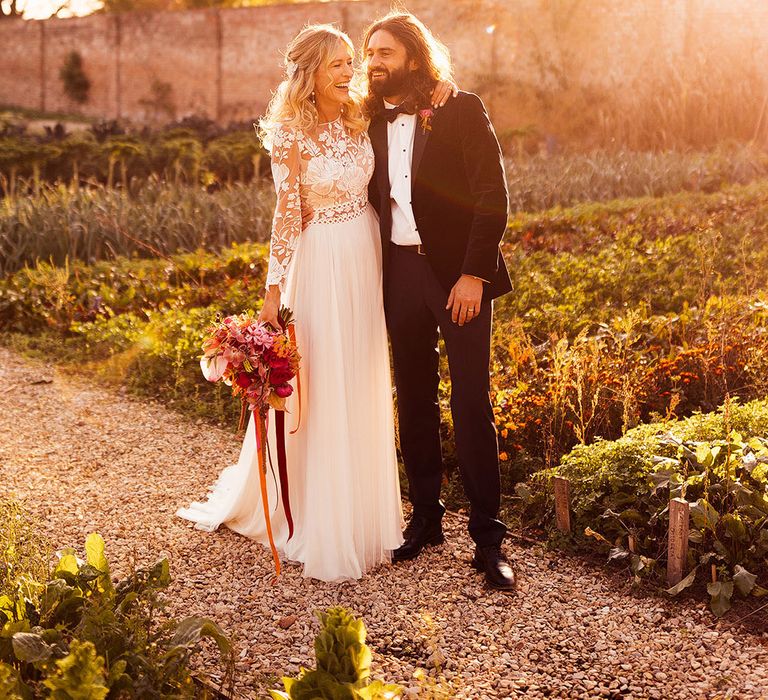 Bride in floral lace wedding dress by Margaux Tardits smiling groom in black tie at golden hour 