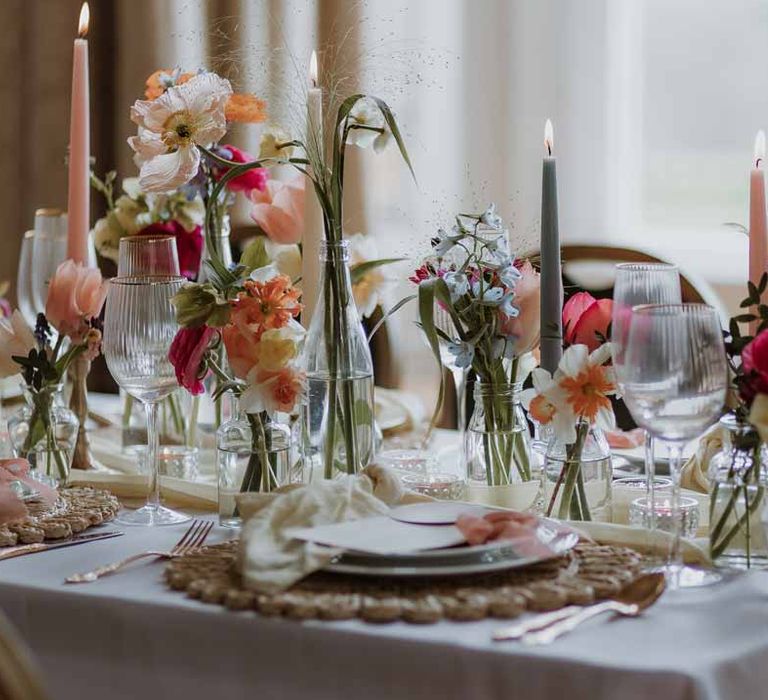 Spring and pastel wedding tablescape with orange poppies, pink and red roses, jasmine and bluebells on white wedding tablecloth and off-white satin table runner with pastel tapered candles, gold-rimmed wine glasses and classic crockery 