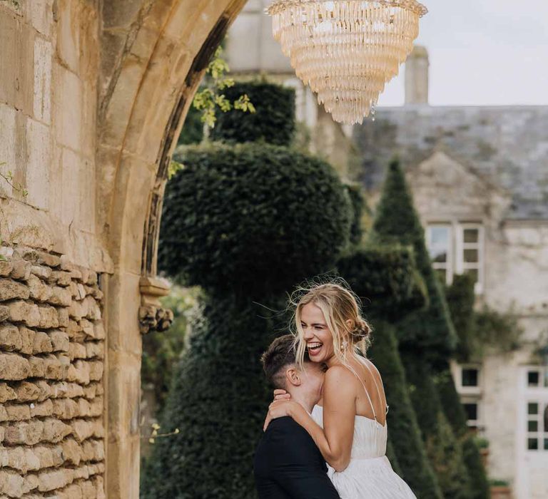 Groom in classic black tuxedo picking up bride in Watters strappy sleeveless princess wedding dress and chapel length wedding veil in archway at Euridge Manor 