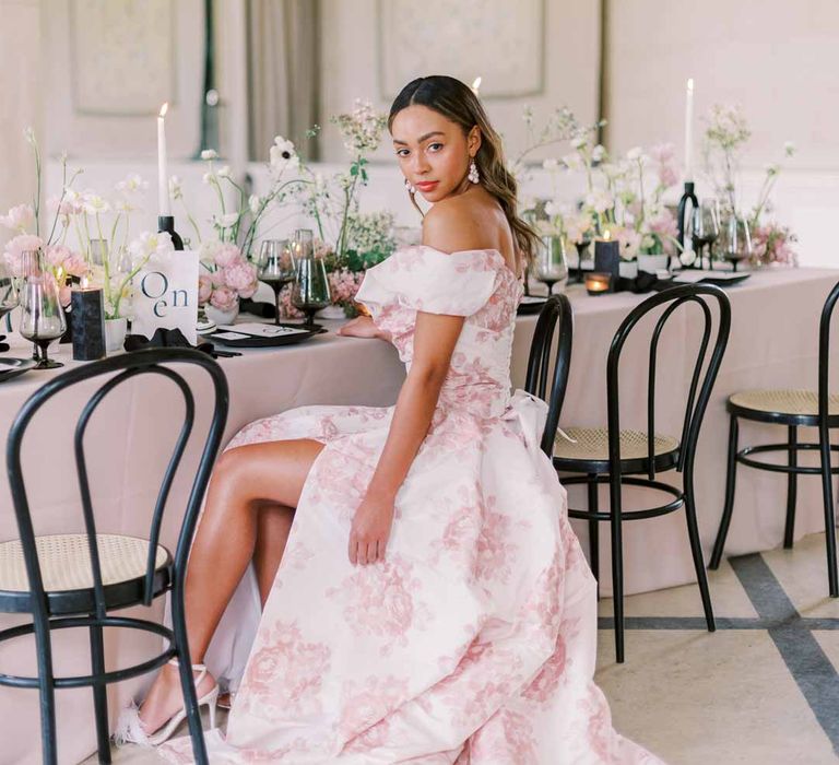 Bride wearing off the shoulder white and pink floral patterned bridal gown with black bow and front slit sitting at pink and monochrome wedding tablescape with light pink wedding tablecloth, white poppies, light pink Persian buttercups, baby's-breath and foliage centrepieces, black artistic candlestick holders, white tapered candles, black crockery and monochrome wedding stationery 