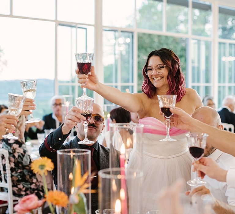 bride with wavy bob hair and glasses wearing a strapless wedding dress toasting with her wedding guests during the wedding breakfast