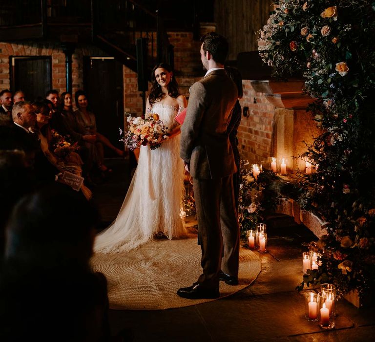 Bride in strapless ivory fringed crepe gown and chapel length veil holding mixed bridal bouquet with garden roses, carnations, peonies and foliage at the alter with groom in chocolate brown grooms suit at Willow Marsh Farm Loughborough 