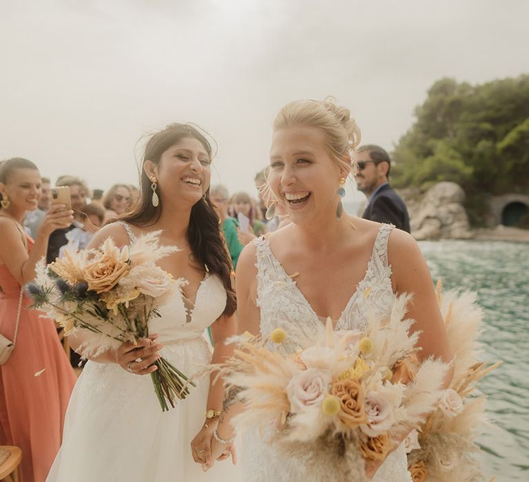 Indian bride and Swiss bride in lace wedding dresses holding dried flower wedding bouquets laughing during their wedding ceremony 