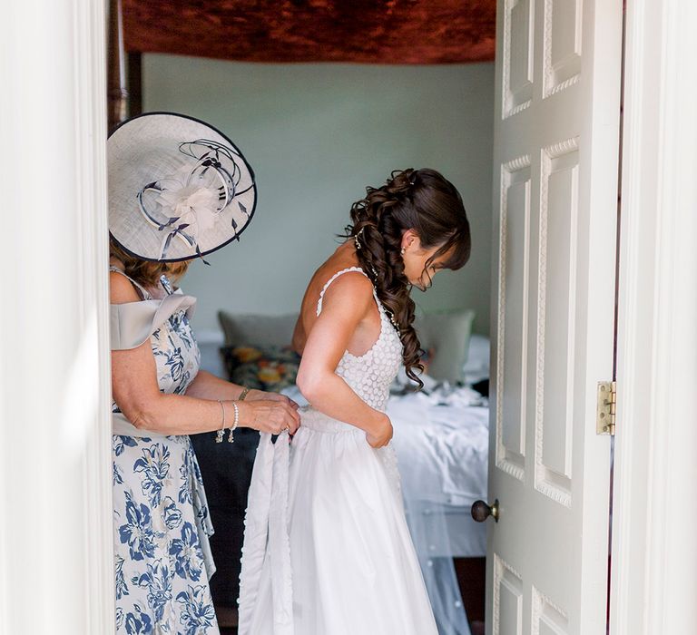 mother of the bride in a blue and white dress and hat helping her daughter into her Made With Love bridal gown on the morning of the wedding 