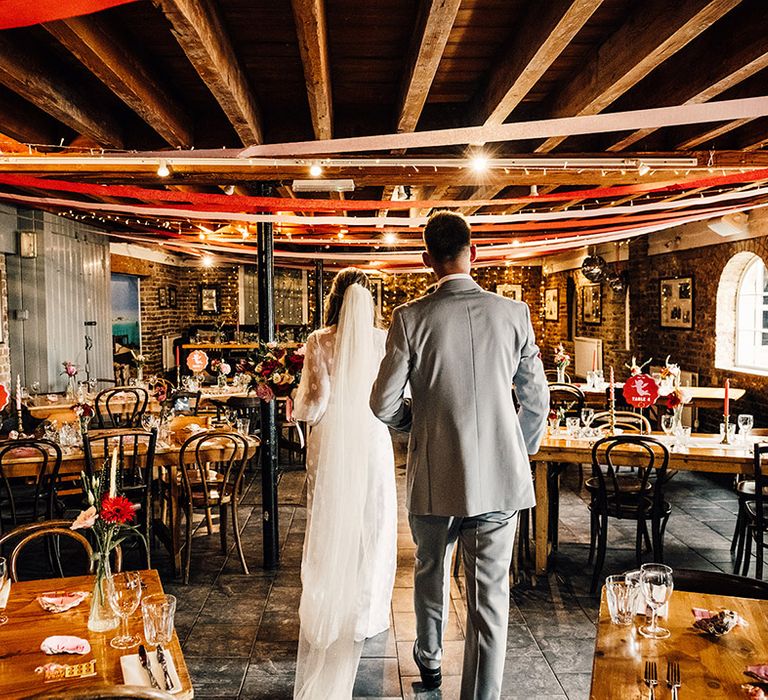 bride in a jumpsuit and groom in a grey suit entering their wedding reception with streamer ceiling decor at East Quay wedding venue 