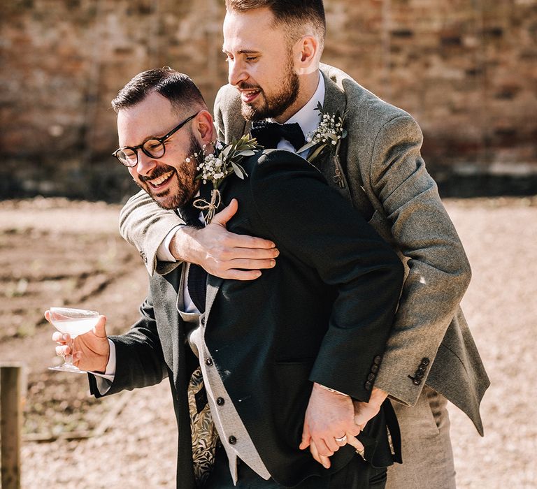 The groom in grey tweed suit embraces groom in black suit for cute wedding photo 