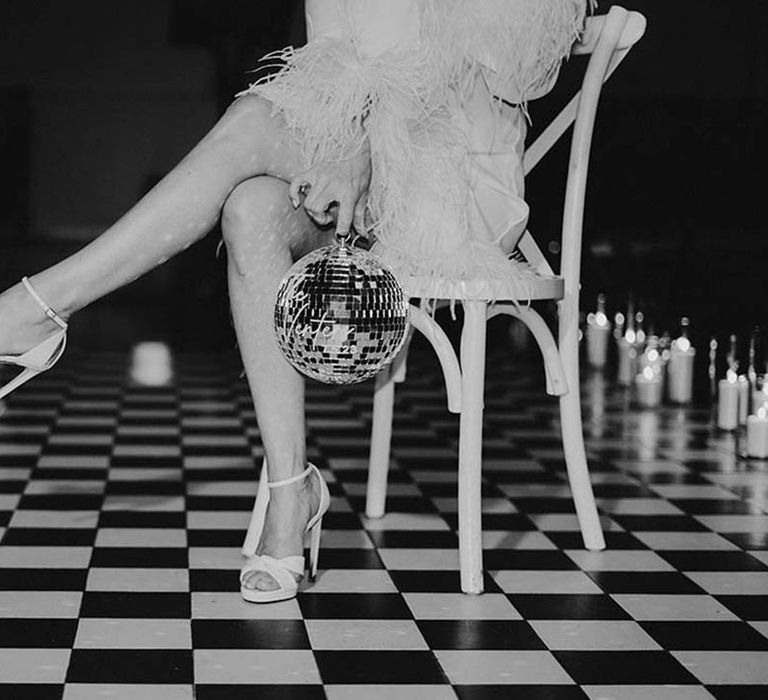 Bride sits on chair showing off white peep toe wedding shoes on checkerboard dance floor with disco balls 