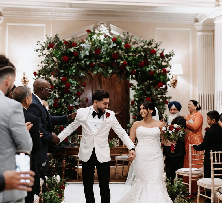 Groom in white tuxedo walks down the aisle with the bride in a lace Enzoani mermaid wedding dress with red rose flowers 