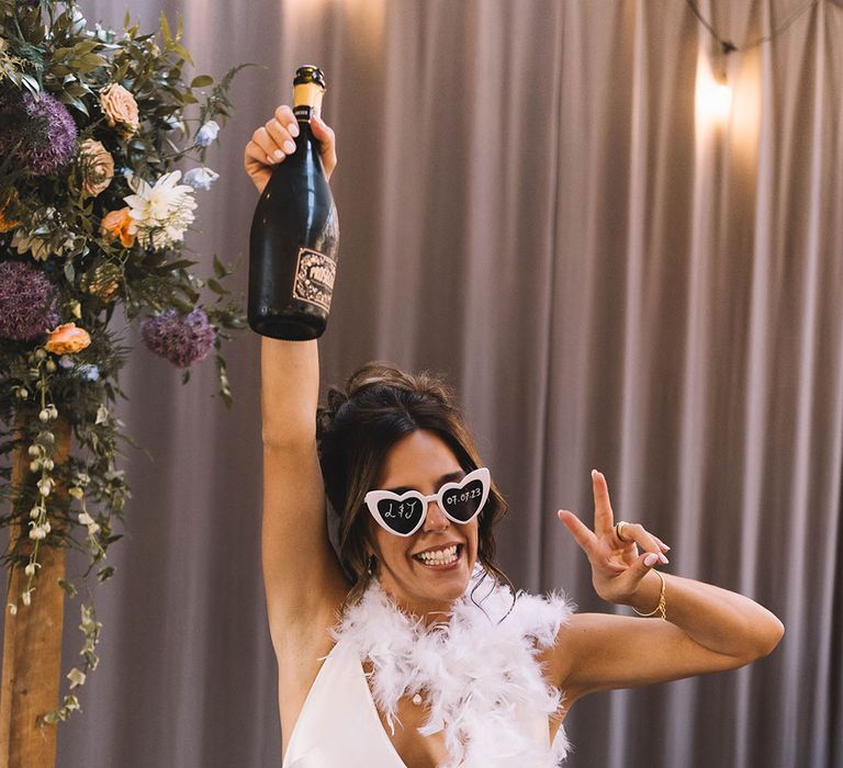 Bride in satin slip wedding dress with white feather boa wearing retro sunglasses and champagne bottle 