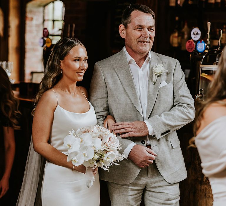 Father of the bride in grey wedding suit with bride in satin slip wedding dress at wedding ceremony 