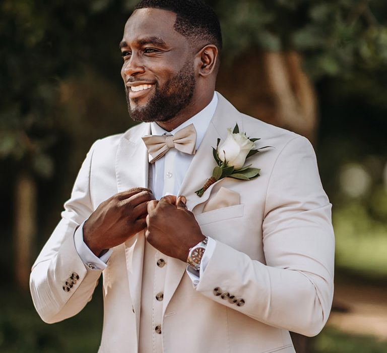 Groom wearing cream wedding three piece suit with white rose buttonhole and champagne bow tie and pocket square 