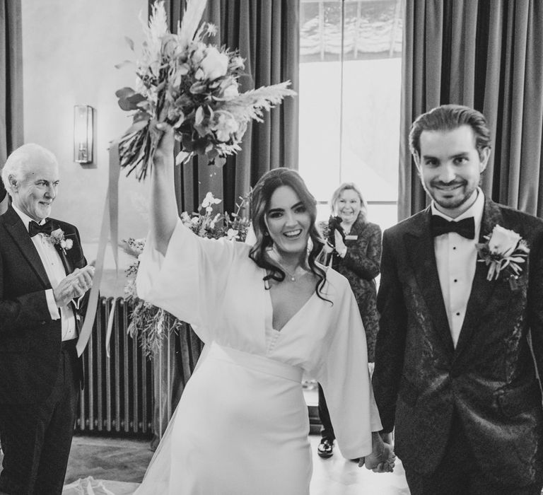 Black and white wedding ceremony photography bride and groom holding hands with the bride celebrating holding her bouquet up and wedding guests in the background