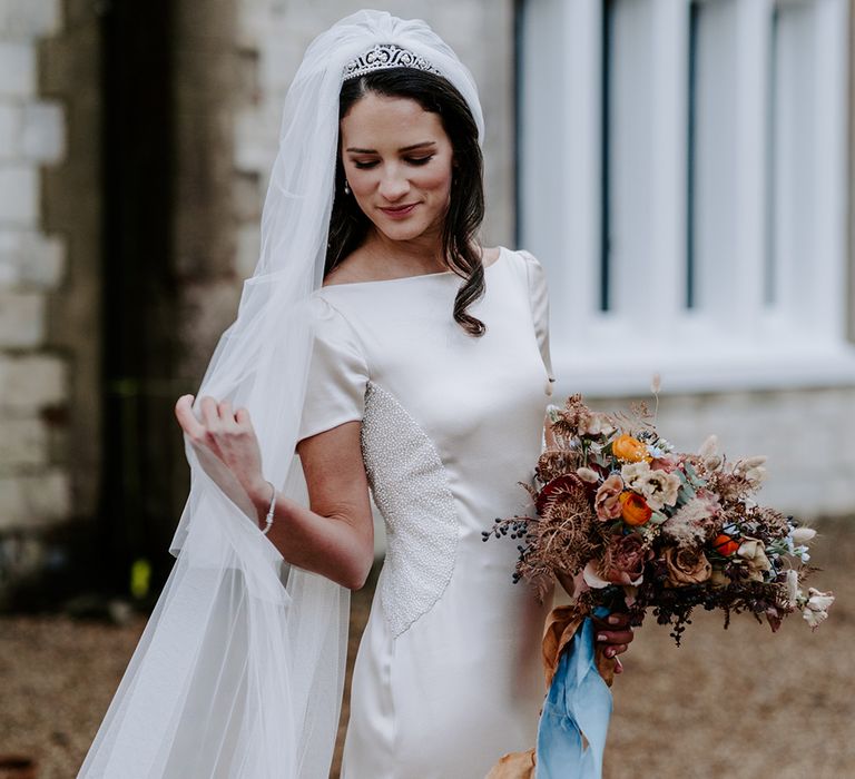 A bride moves her veil over her shoulder as she smiles down to the ground. She holds a winter wedding bouquet in her hand that is ties with a blue ribbon