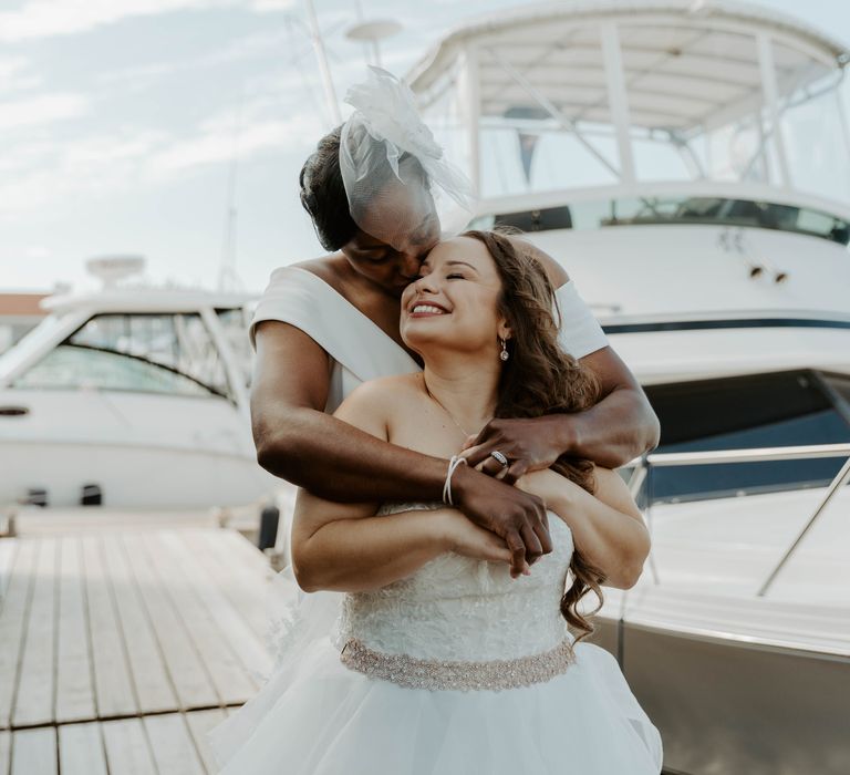 Two brides embracing on the marina dock with a boat in the background 