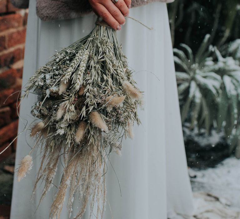 Bride in fur coat holds artisan dried flower bouquet whilst stood in gardens