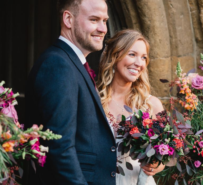 Bride & groom stand and smile outside the church surrounded by bright flowers