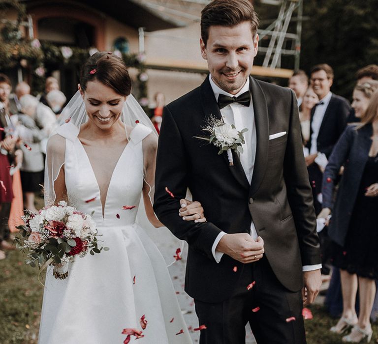 Smiling bride in white deep V neck Jesus Piero wedding gown and groom in black Tiger of Sweden tuxedo walk arm-in-arm through confetti at German wedding