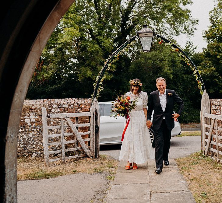 Bride in a white Broderie Anglaise wedding dress with colourful embroidery pattern arriving at the church with her father 