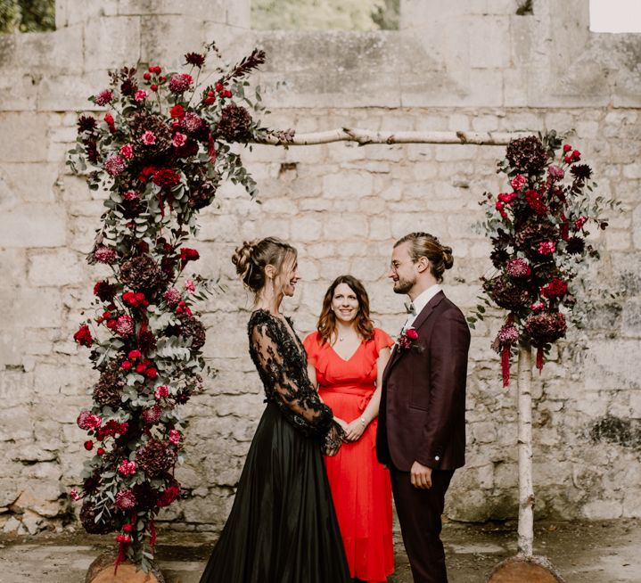 Bride and groom standing at the altar in a black dress and burgundy suit exchanging vows 