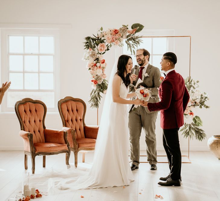 Groom in a burgundy tuxedo and bride in a fitted wedding dress laughing at their Core Clapton wedding ceremony 