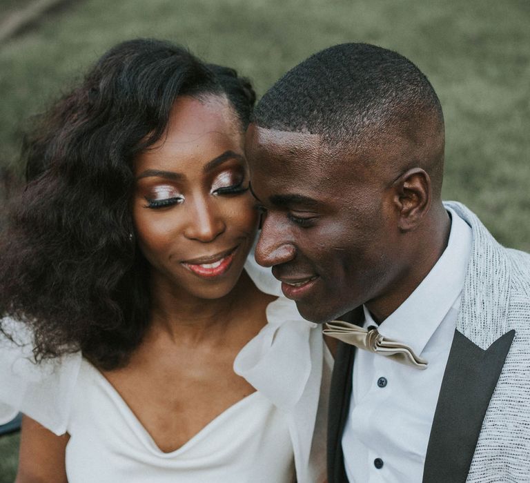 Smiling bride with curled hair in white satin dress and groom in grey suit jacket with satin bow tie sit together on bench outside after Bridge Community Church wedding