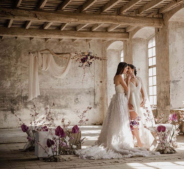 Two brides kissing at romantic ceremony at industrial wedding venue with purple flowers and drift wood hanging installation in a ruffle skirt mini wedding dress