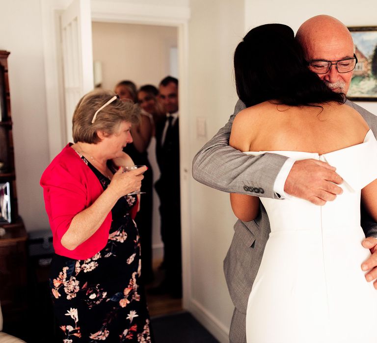 Bride in strapless Vagabond wedding dress hugs father in grey suit whilst wedding dress in floral dress and pink jacket stands in the background