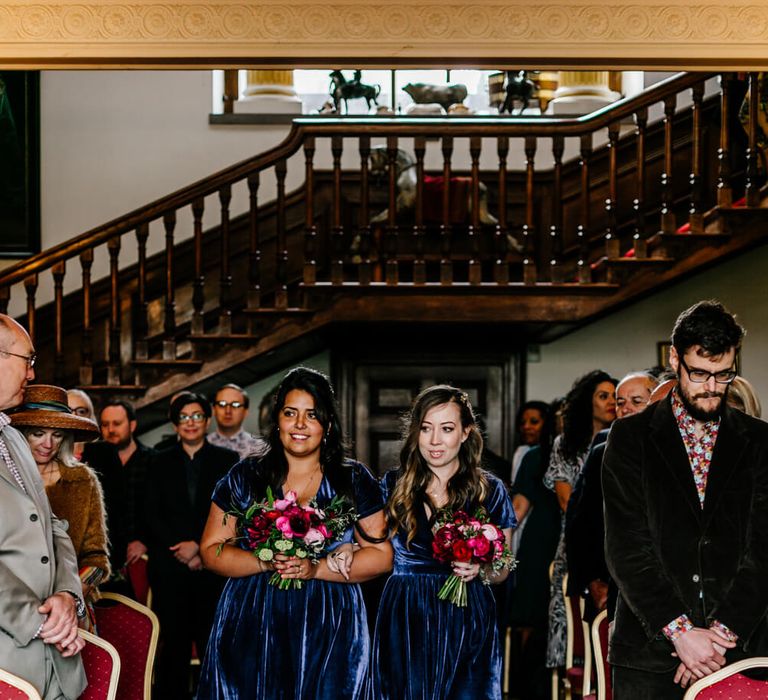 Two bridesmaids walking down the aisle wearing knee length velvet navy bridesmaid dresses with red shoes and red bouquets | Lindy Bop Dresses | Photography by Recommended Supplier EPIC LOVE STORY