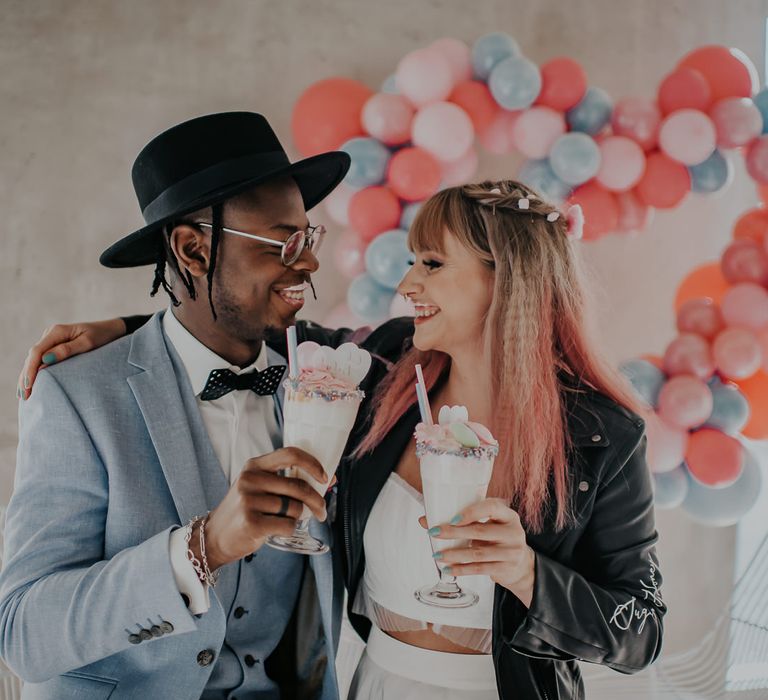 Bride in a leather jacket and groom in a blue suit enjoying a milkshake together topped with pastel flying saucers