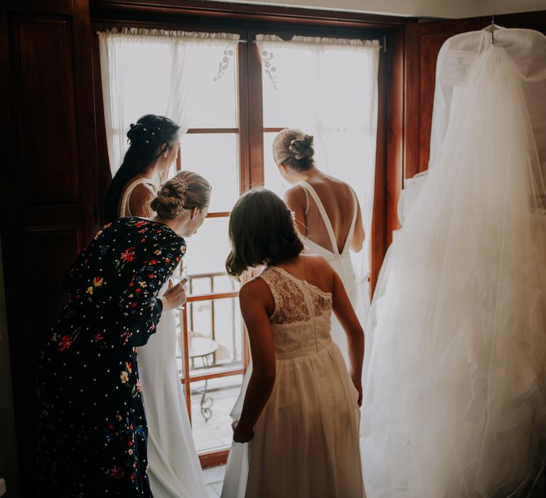 The bride and bridesmaids getting ready and looking out the window, her dressing hanging up beside them