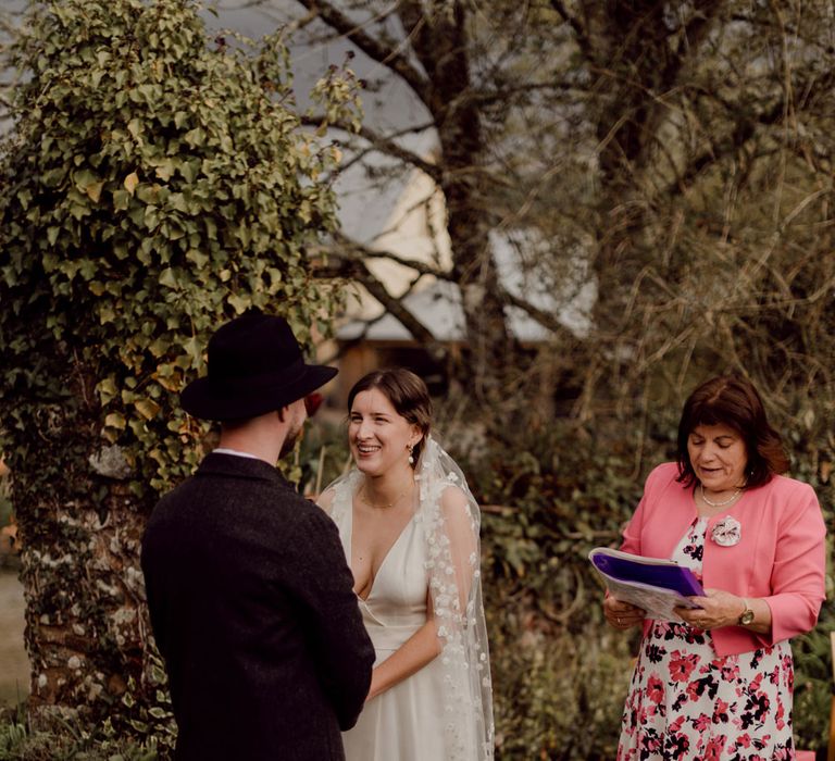 Bride in Charlie Brear wedding dress and daisy chapel length applique veil smiles whilst holding hands with groom in grey tweed suit and black fedora at garden party wedding at Hayne, Devon