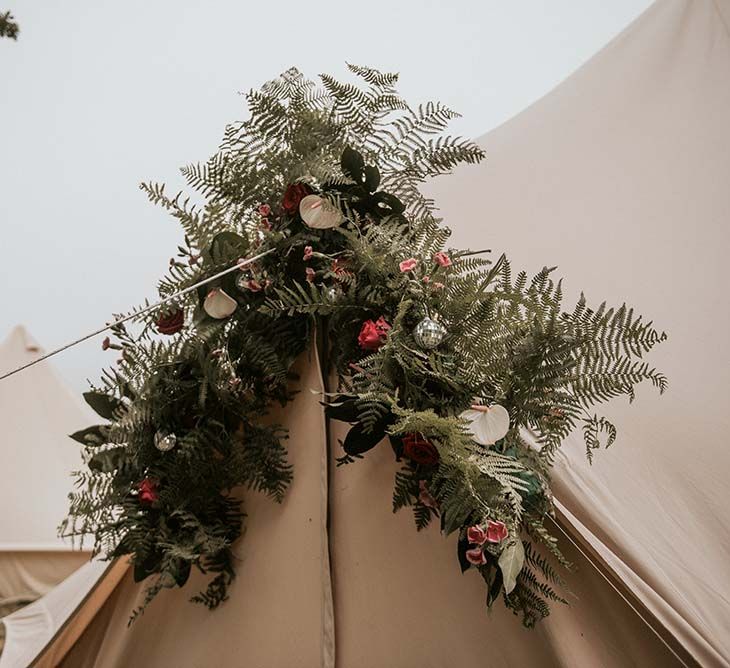 Fern, mini disco ball and tropical flower installation at teepee entrance for festival themed wedding