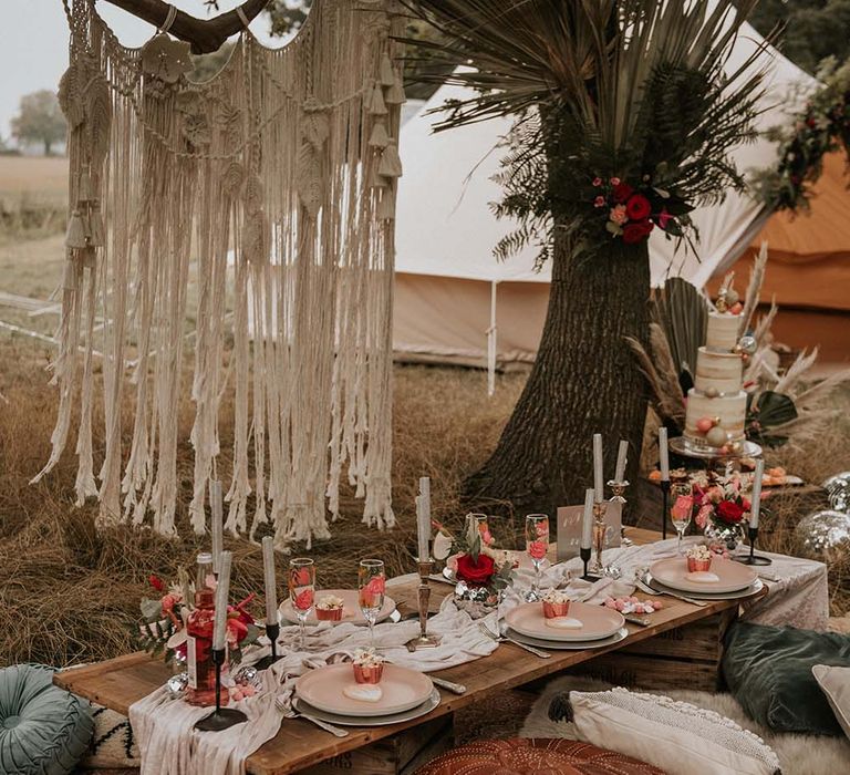 Low wooden wedding tablescape with beige table runner, long silver candles, pink pastel colours and macrame hanging outside at festival themed wedding