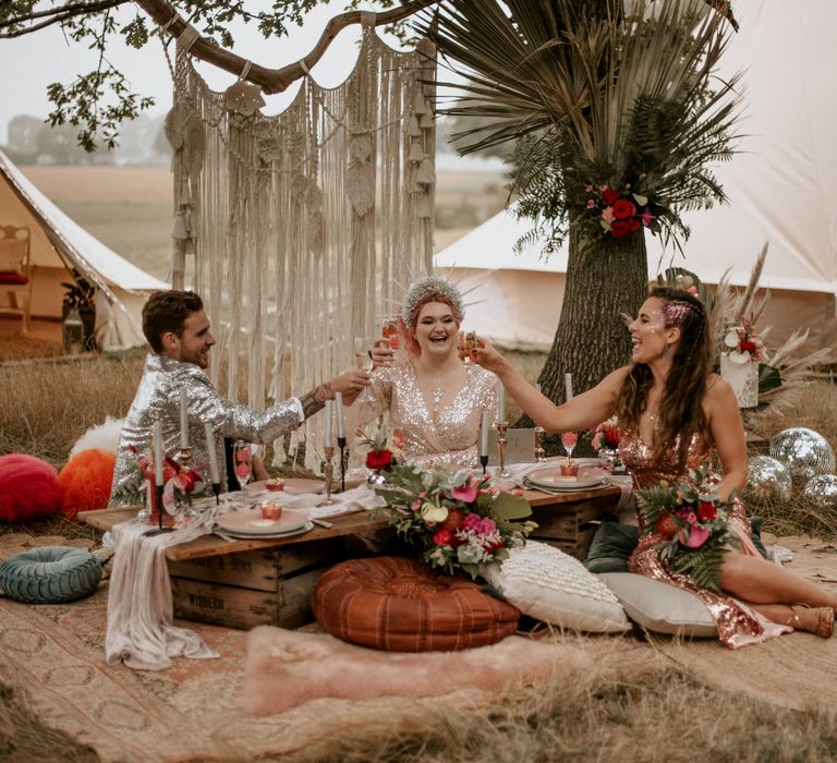 Two brides in sequin dresses and face gens sit at low wooden table outside raising their glass to groom in silver sequin suit jacket at festival themed wedding