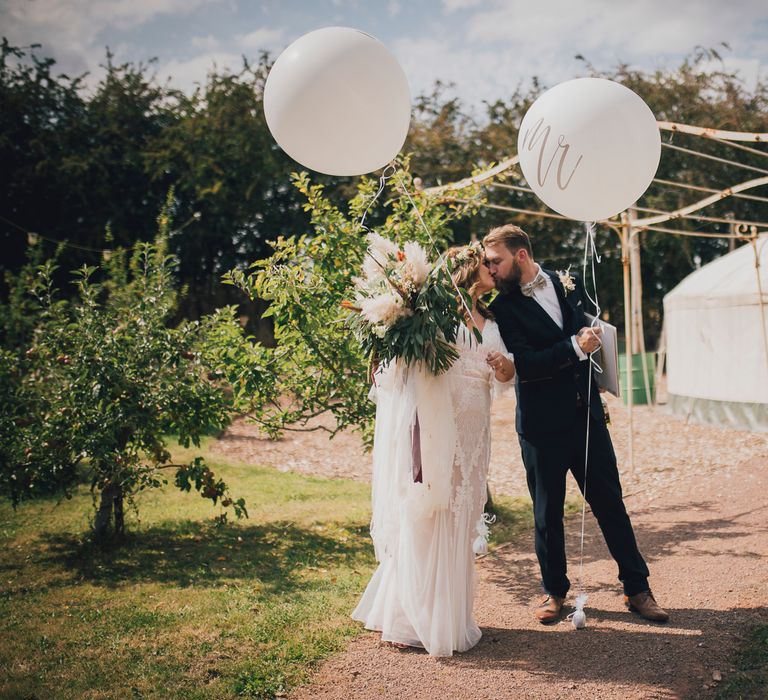 Bride & groom stand with white balloons outside yurt 