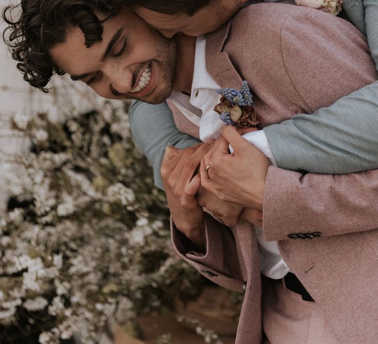 Grooms embrace whilst whilst wearing floral buttonholes 