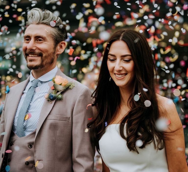 Bride in white strapless Rebecca Vallance Dress holds hands with groom in brown Moss Bros suit and blue tie as they walk through multicoloured confetti at the Hotel du Vin after Harrogate wedding