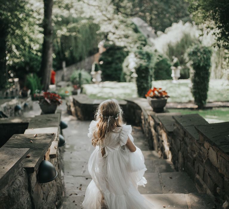 A flower girl in a pleated long white dress runs down steps.