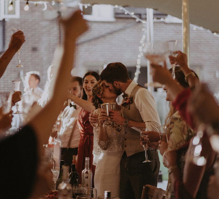 Guests all raise a glass to bride in white Rime Arodaky dress and groom in light brown waistcoat who kiss in tipi during wedding breakfast at garden wedding with burnt orange wedding theme