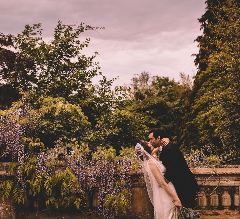 Groom in a black wedding suit kissing his bride in a satin wedding dress and Juliet cap wedding veil 