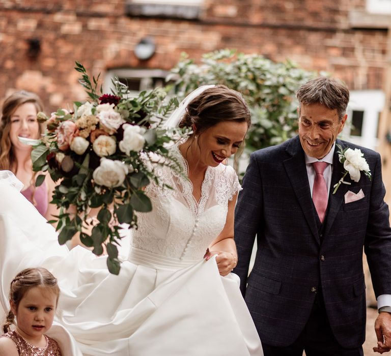 Bride in lace cap sleeved wedding dress holding white, red and green bridal bouquet walks through the courtyard at The West Mill Derby with man in checked suit and pink tie before wedding