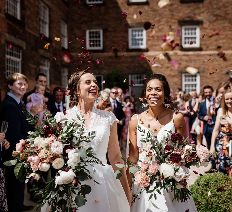 Smiling bride in white cami wedding dress holds hands with bride in white lace capped sleeve wedding dress as they both carry white, pink and green bridal bouquets outside The West Mill Derby whilst guests shower them with multicoloured confetti