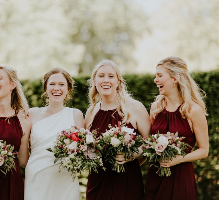 Bride in a one shoulder sateen wedding dress laughing with her bridesmaids in halter neck burgundy dresses holding their bouquets 