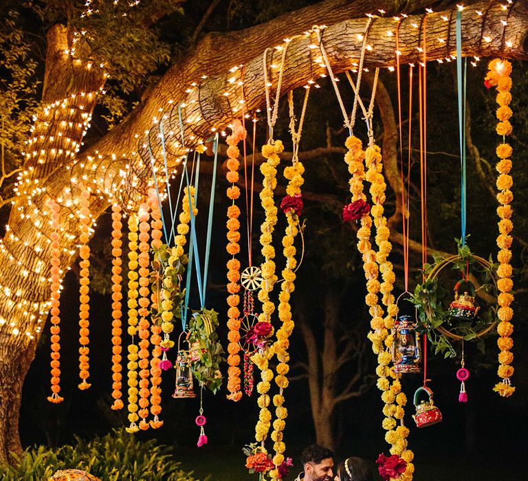 An Indian couple sit on a swing at their multicultural wedding. The swing is covered in yellow flowers. She wears a gold dress and he wears a blue suit.