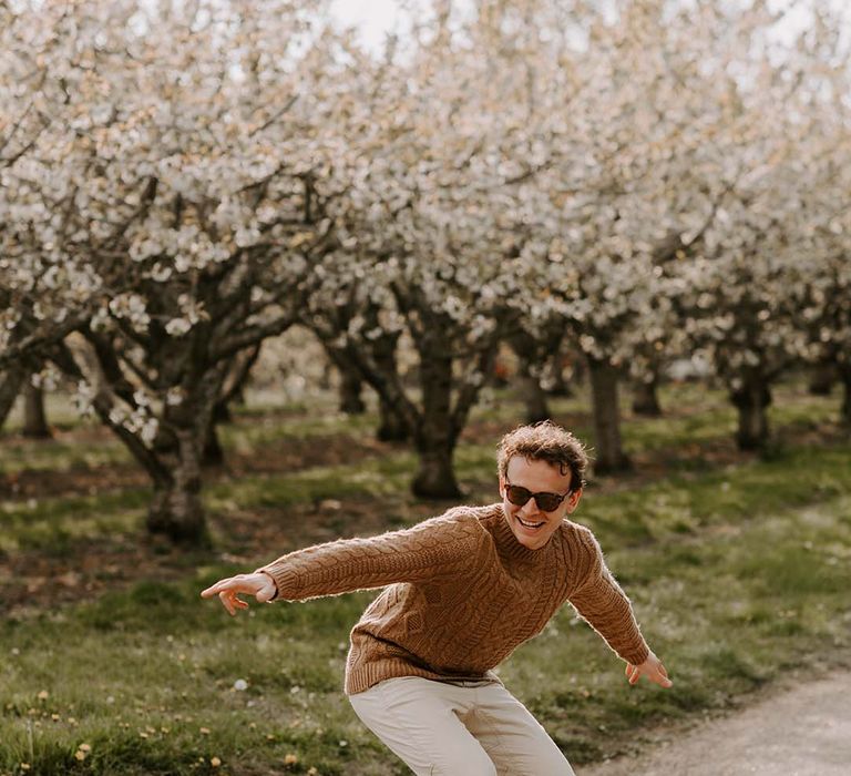 Groom with curly hair riding a skateboard in trainers, chinos, cable knit jumper and shades 