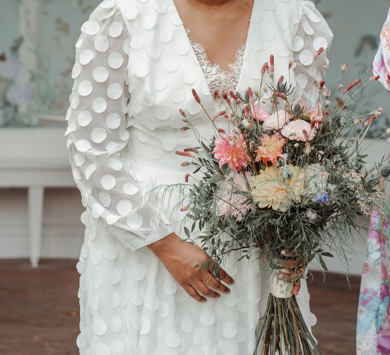 Laughing bride in white long mesh sleeve spotted Vagabond wedding dress and bridal hair bow holding mixed bridal bouquet stands outside Victorian Summerhouse at Wasing Park wedding