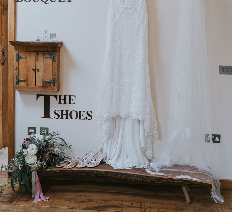 Lace wedding dress with train hung next to veil with lace trim, white heeled sandals, mixed bridal bouquet and sandals before Tythe Barn wedding with barn wedding flowers